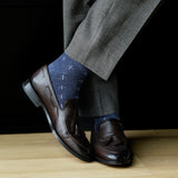 Brown loafers with tassels worn with gray pants and blue patterned socks on a wooden floor.