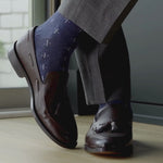 Brown leather loafers with tassels worn with blue patterned socks and gray pants on a wooden floor.