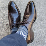 Man sitting with legs crossed wearing gray, white and blue stripe socks with brown shoes