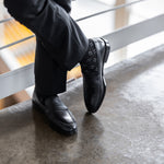 Model wearing black and grey flower-petal dress socks with black trousers and black leather penny loafers, standing crossed-leg view.