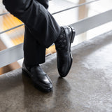 Model wearing black and grey flower-petal dress socks with black trousers and black leather penny loafers, standing crossed-leg view.