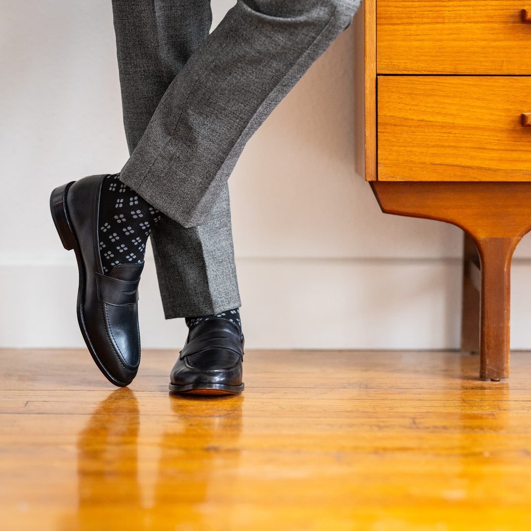 Model wearing black and grey flower-pattern dress socks with grey trousers and black leather penny loafers, standing crossed-leg view.