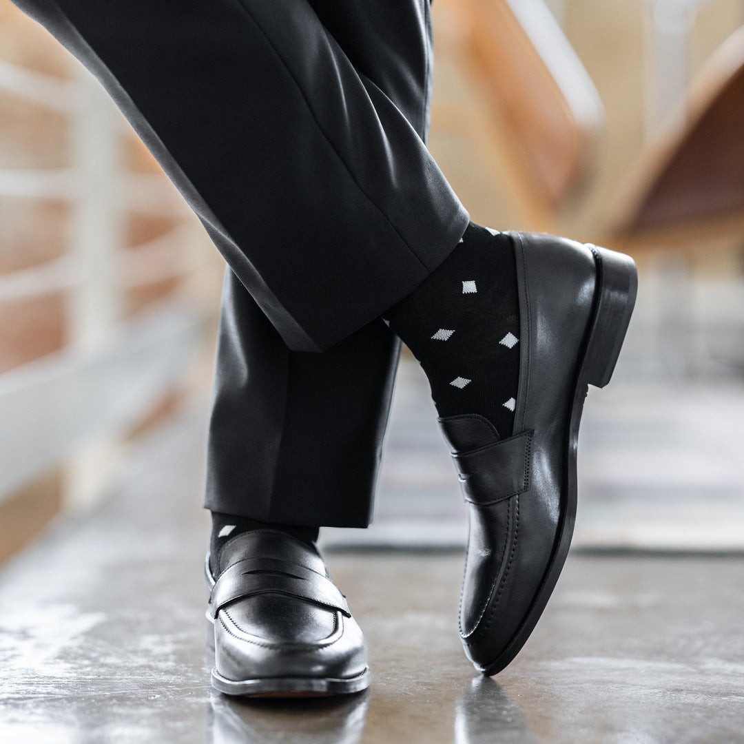Model wearing black and grey micro-diamond dress socks with black trousers and black leather penny loafers, standing crossed-leg view.