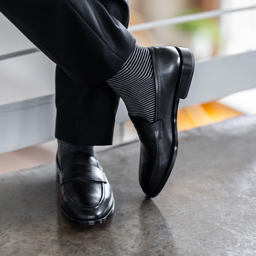 Model wearing black and grey striped dress socks with black trousers and black leather penny loafers, standing crossed-leg view.