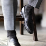 The Black & Silver Square-Grid dress socks with tailored trousers and loafers in a seated lounge view