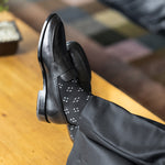 Model wearing black and grey flower-petal dress socks with black trousers and black leather penny loafers, reclined feet-up view.