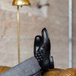 Model wearing black and grey flower-pattern dress socks with grey trousers and black leather penny loafers, reclined feet-up view.
