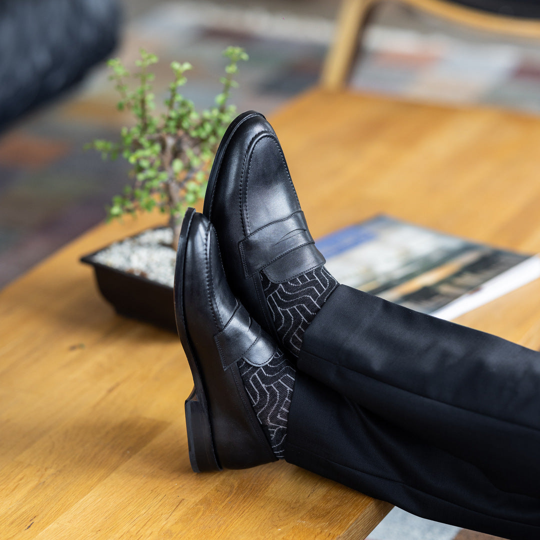 Model wearing black and grey geometric-maze dress socks with black trousers and black leather penny loafers, reclined feet-up view.
