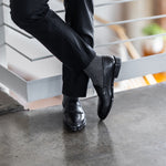 Model wearing black and grey striped dress socks with black trousers and black leather penny loafers, standing crossed-leg view.