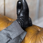 Model wearing black and grey diamond-grid dress socks with grey trousers and black leather penny loafers, reclined feet-up view.