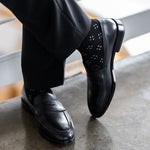 Model wearing black and grey flower-petal dress socks with black trousers and black leather penny loafers, standing crossed-leg view.