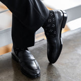Model wearing black and grey flower-petal dress socks with black trousers and black leather penny loafers, standing crossed-leg view.