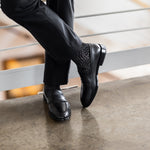 Model wearing black and grey geometric-maze dress socks with black trousers and black leather penny loafers, standing crossed-leg view.