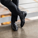 Model wearing black and grey geometric-maze dress socks with black trousers and black leather penny loafers, standing crossed-leg view.