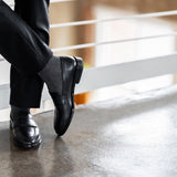 Model wearing black and grey striped dress socks with black trousers and black leather penny loafers, standing crossed-leg view.
