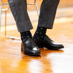 Model wearing black and grey diamond-grid dress socks with grey trousers and black leather penny loafers, standing crossed-leg view.