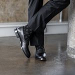 Model wearing black and grey flower-petal dress socks with black trousers and black leather penny loafers, standing crossed-leg view.