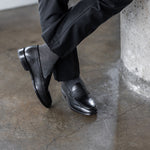 Model wearing black and grey striped dress socks with black trousers and black leather penny loafers, standing crossed-leg view.