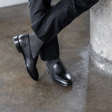 Model wearing black and grey striped dress socks with black trousers and black leather penny loafers, standing crossed-leg view.