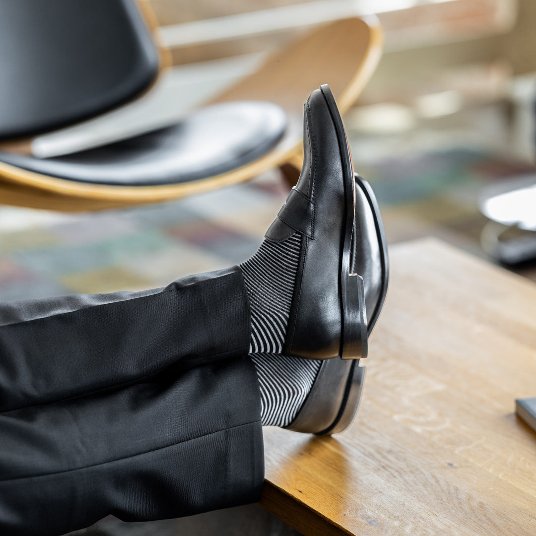 Model wearing black and grey geometric-maze dress socks with black trousers and black leather penny loafers, reclined feet-up view.
