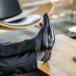 Model wearing black and grey geometric-maze dress socks with black trousers and black leather penny loafers, reclined feet-up view.