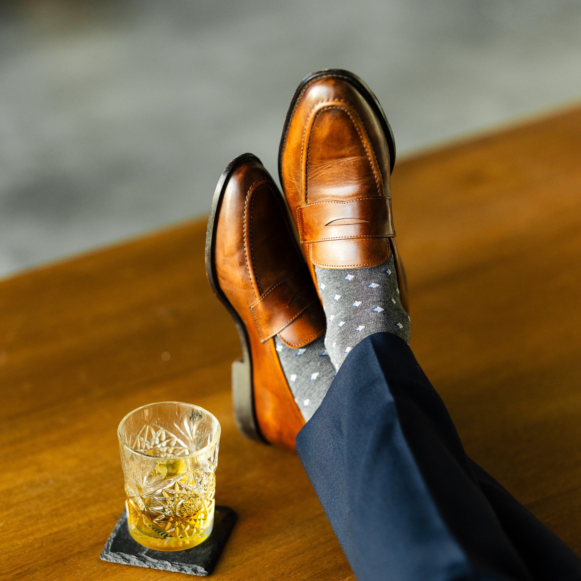Grey Gemstones dress socks with navy trousers and brown penny loafers beside a whiskey glass