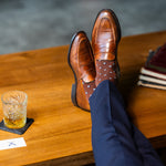 Brown Gemstones dress socks with navy trousers and brown penny loafers beside a whiskey glass