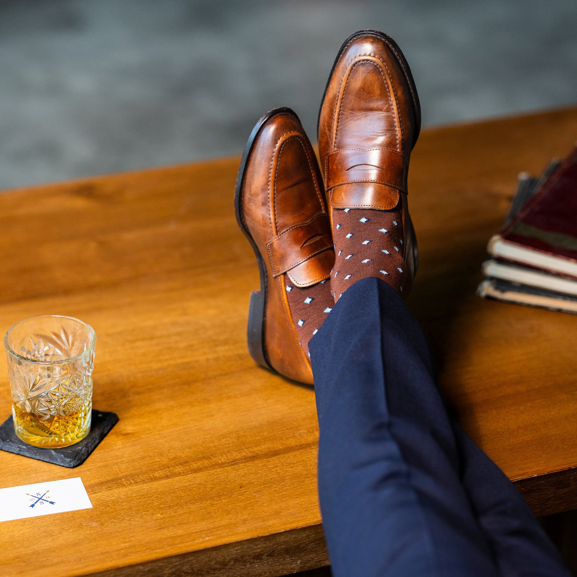 Brown Gemstones dress socks with navy trousers and brown penny loafers beside a whiskey glass