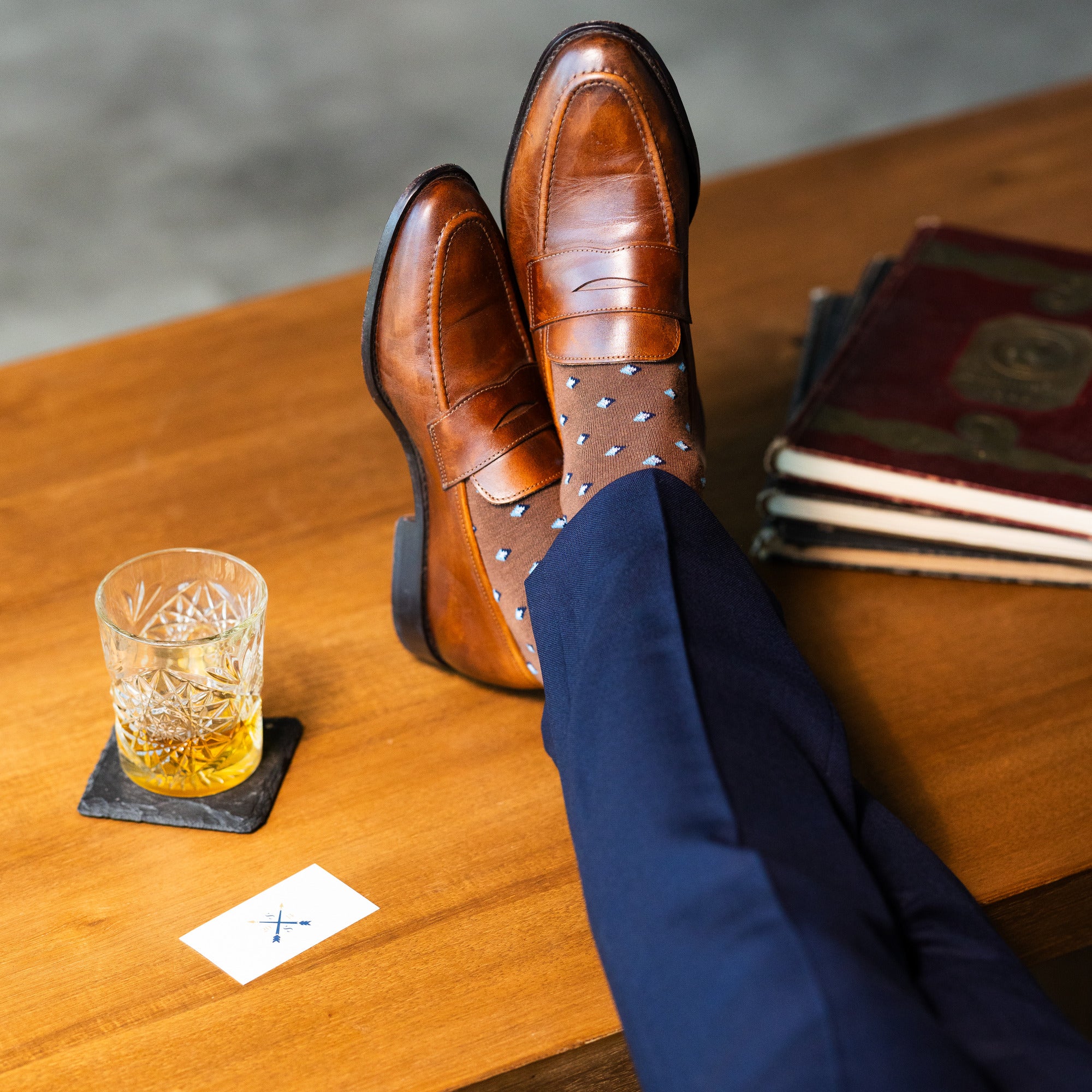 Brown Gemstones dress socks with navy trousers and loafers beside a whiskey glass