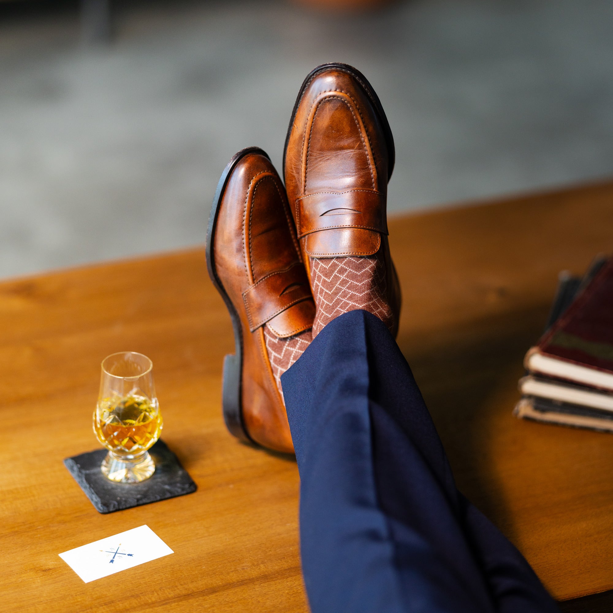 Brown patterned dress socks with navy trousers and brown penny loafers beside a whiskey glass