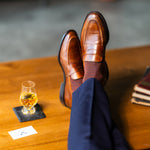 Brown patterned dress socks with navy trousers and brown penny loafers beside a glass