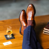 Brown patterned dress socks with navy trousers and brown penny loafers beside a glass