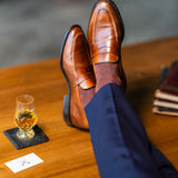Brown patterned dress socks with navy trousers and penny loafers beside a whiskey glass