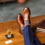 Brown patterned dress socks with navy trousers and loafers beside a whiskey glass