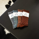 Four folded Gemstones dress socks in Slate Grey, Sage Green, Mocha, and Pecan on a black surface