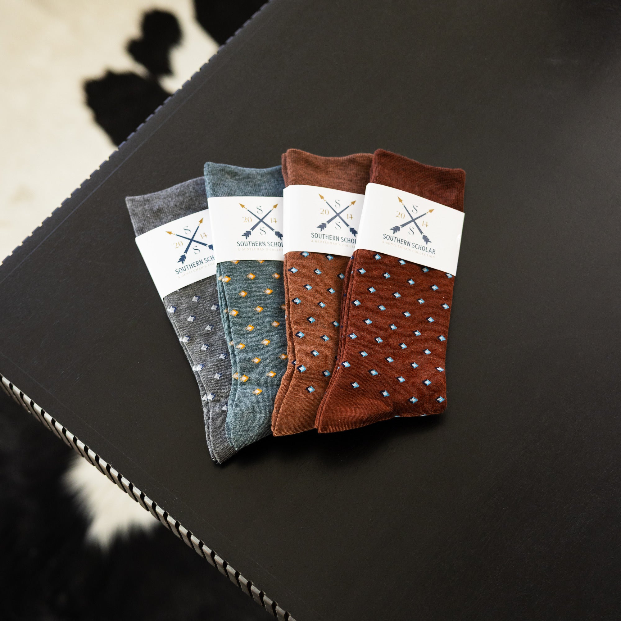 Four folded Gemstones dress socks in Slate Grey, Sage Green, Mocha, and Pecan on a black surface