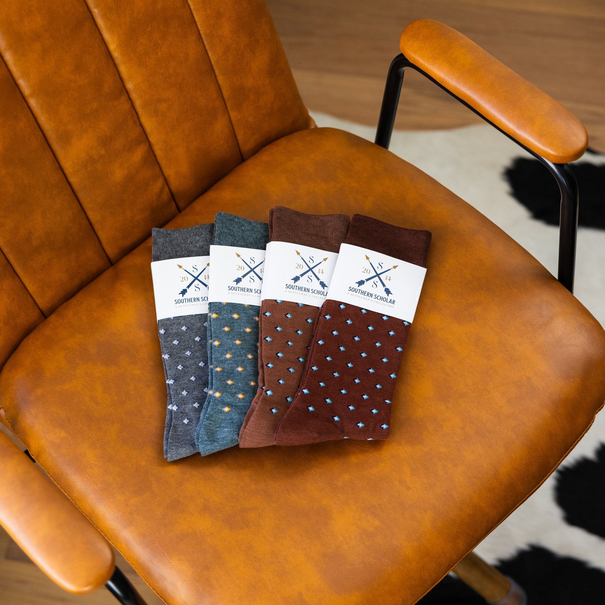 Four folded Gemstones dress socks in Slate Grey, Sage Green, Mocha, and Pecan on a leather chair