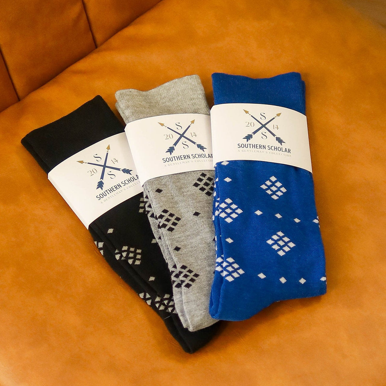 Three pairs of folded socks displayed on an orange-brown leather chair: one black pair with white geometric patterns, one gray pair with white geometric patterns, and one blue pair with white diamond patterns. Each pair has a white paper band with the brand 'Western Socks' and crossed arrows printed on it.