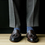 Brown loafers with tassels worn with gray trousers and patterned socks on a wooden floor.