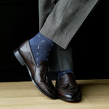 Brown loafers with tassels worn with gray pants and blue patterned socks on a wooden floor.