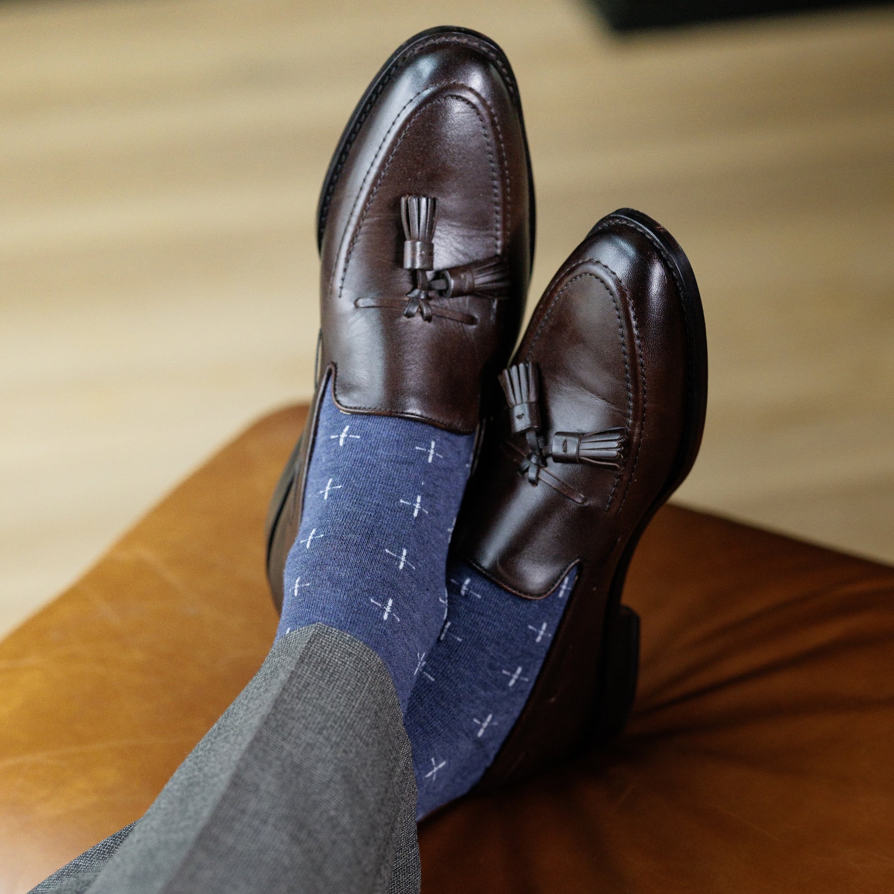 Brown leather loafers with tassels worn with blue patterned socks and gray pants on a wooden floor.