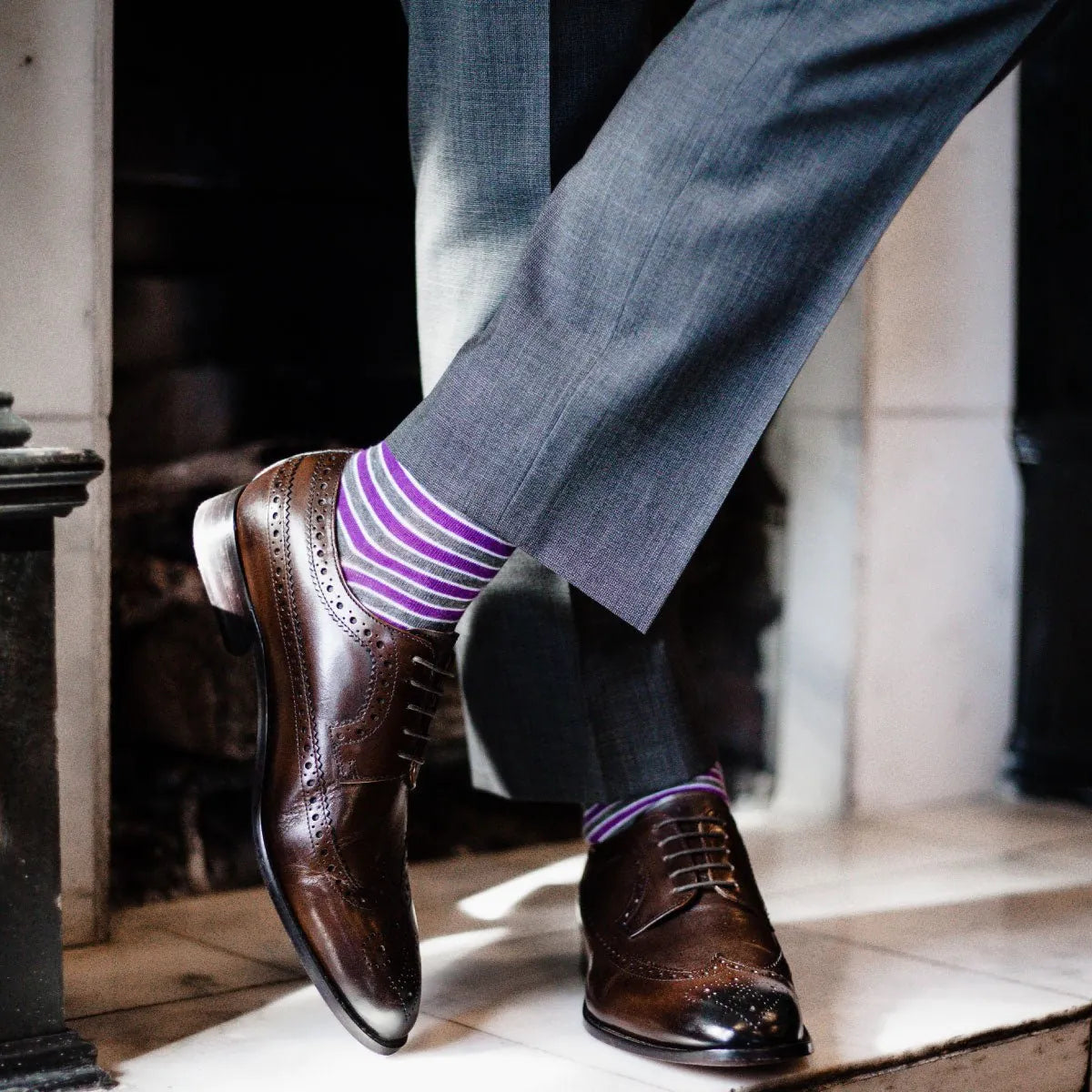 A man wearing purple, grey, and white striped dress socks, brown dress shoes, and grey slacks