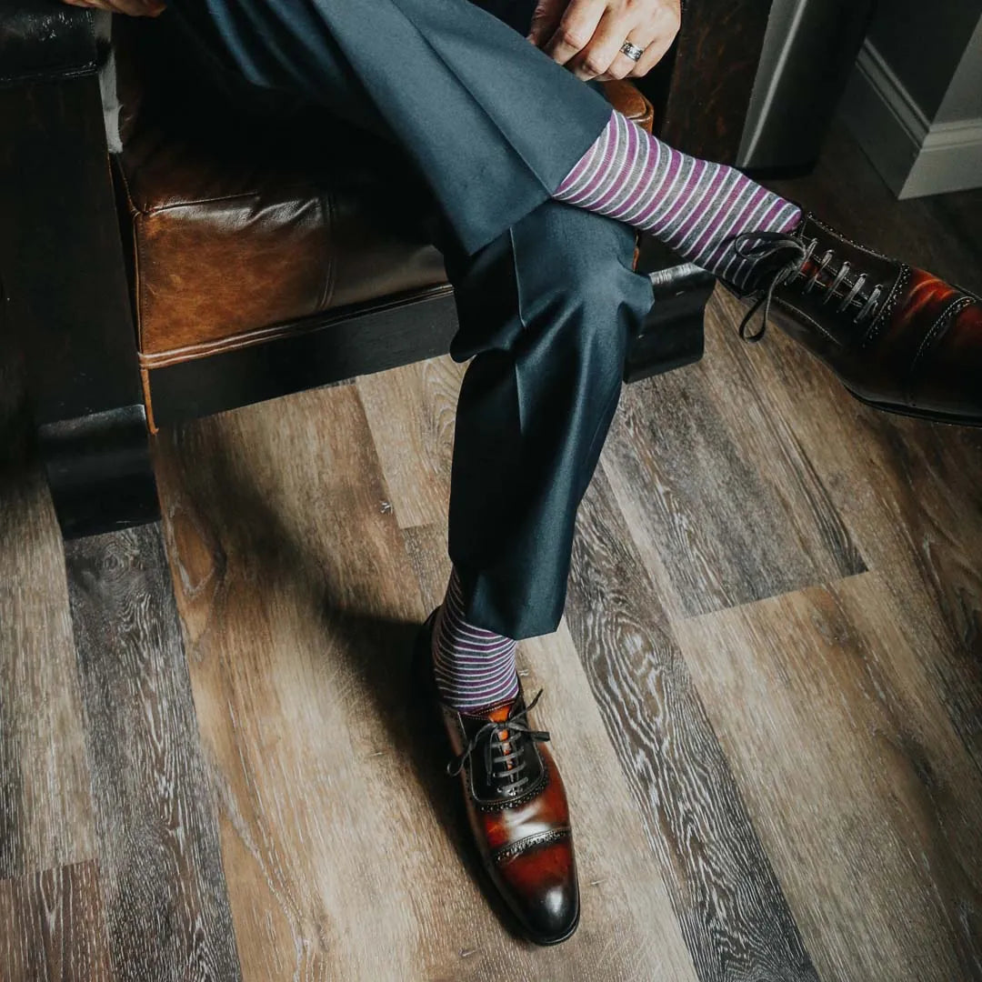 Man sitting with legs crossed wearing purple, grey, and white striped dress socks and brown dress shoes