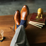 Brown leather shoes with blue polka dot socks on a wooden table with books and a glass.