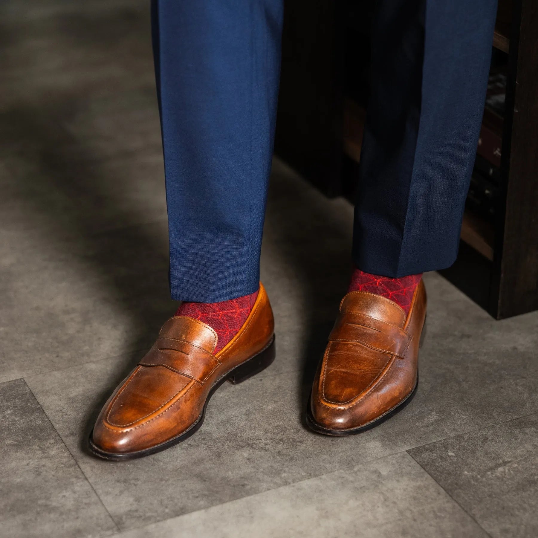 Brown loafers with red patterned socks worn with blue pants on a gray floor.