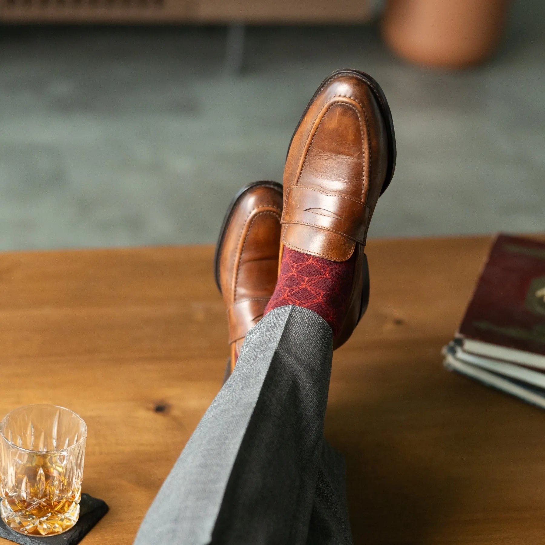 Person wearing brown leather shoes and red patterned socks, with a glass of whiskey on a wooden table.