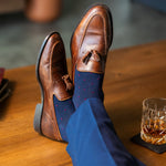 Navy blue socks with a red pattern worn with navy trousers and brown tassel loafers, sitting on a wooden table with a glass of whiskey.
