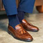 Navy blue socks with a red pattern worn with navy trousers and brown tassel loafers, sitting on a wooden table with a glass of whiskey.