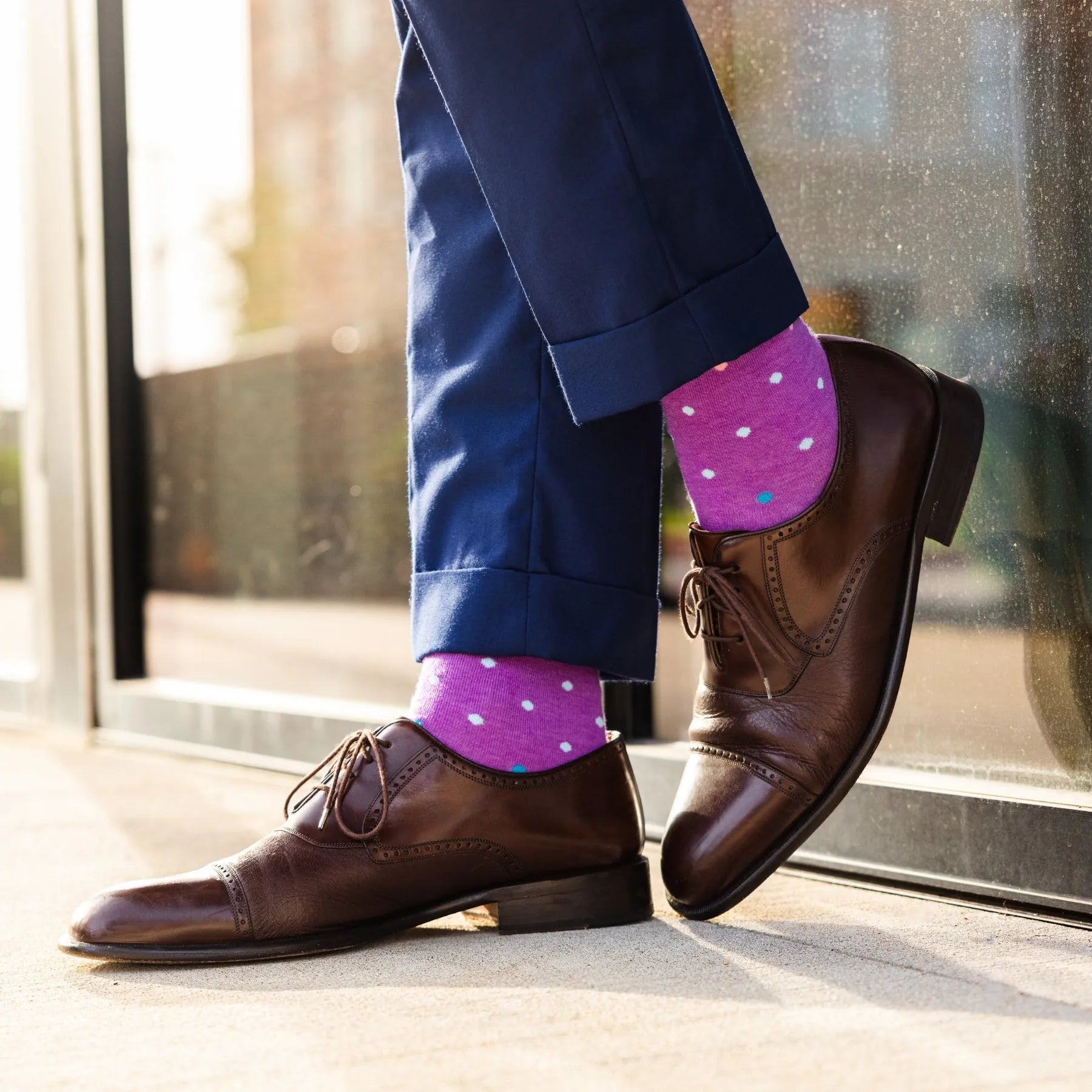 Person wearing purple polka dot socks with brown shoes and blue pants by a window.