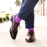 Person wearing brown dress shoes and purple socks with multi-colored polka dots on a blurred outdoor background.